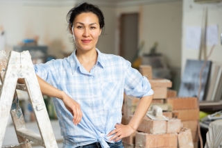 confident smiling young adult woman in casual work clothes, standing next to step ladder surrounded by home improvement materials